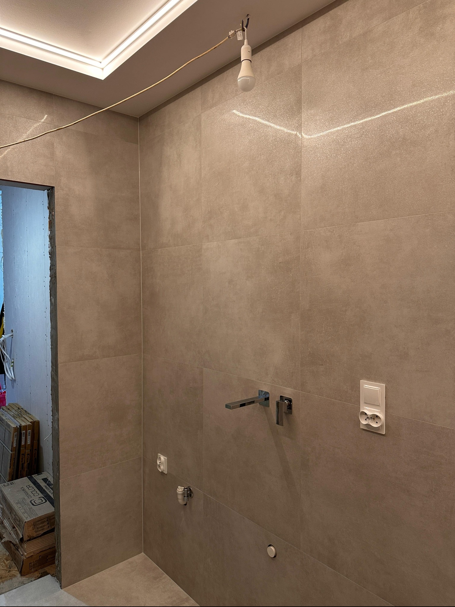 Wykończona ściana łazienki z szarymi płytkami, widoczne przyłącza wody i gniazdko elektryczne. W tle stos płytek i surowy otwór drzwiowy.