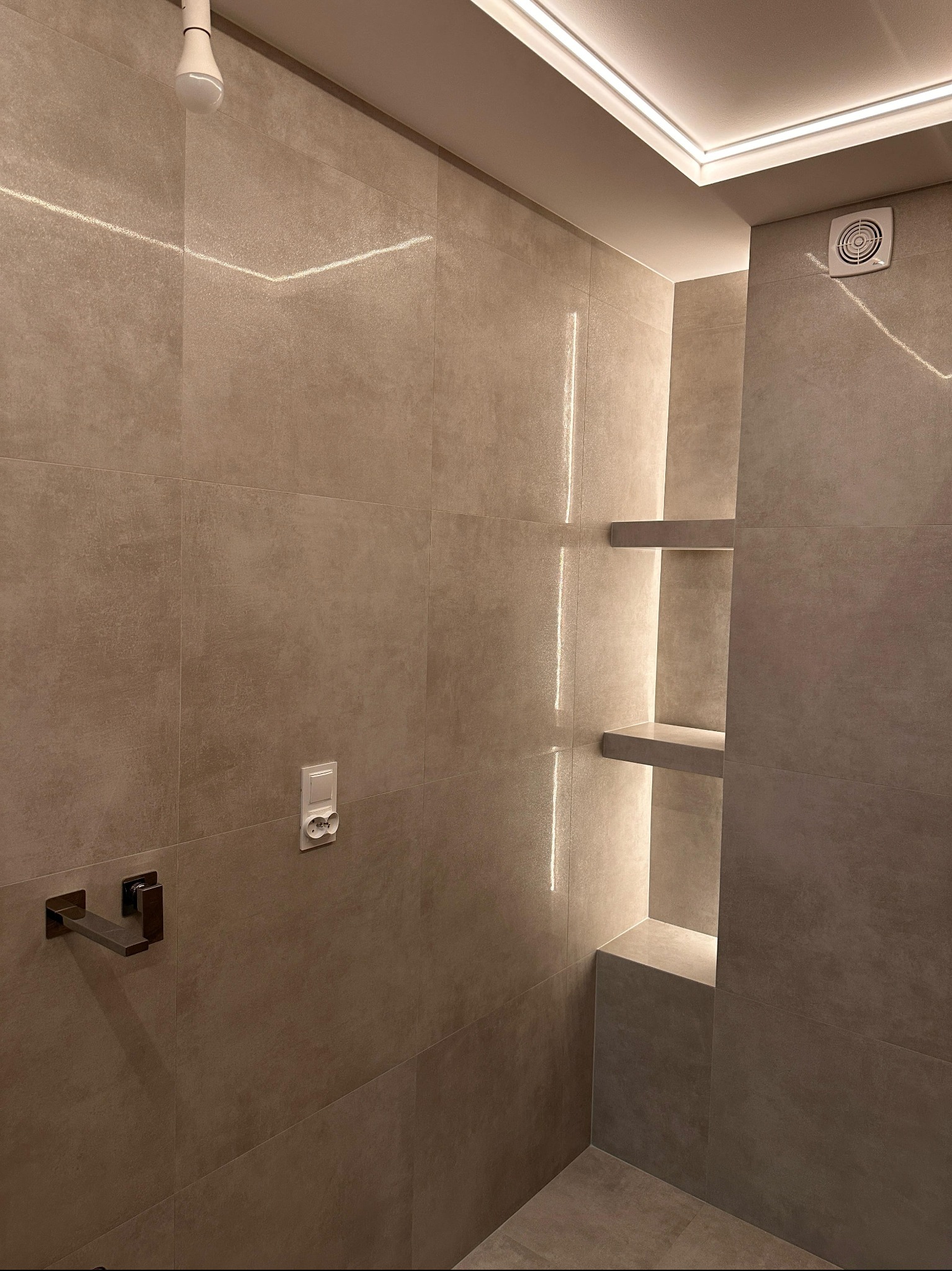 Nowoczesna łazienka z beżowymi płytkami, oświetleniem LED i wnękami z półkami. Minimalistyczny design, widoczny fragment kabiny prysznicowej i wentylator.