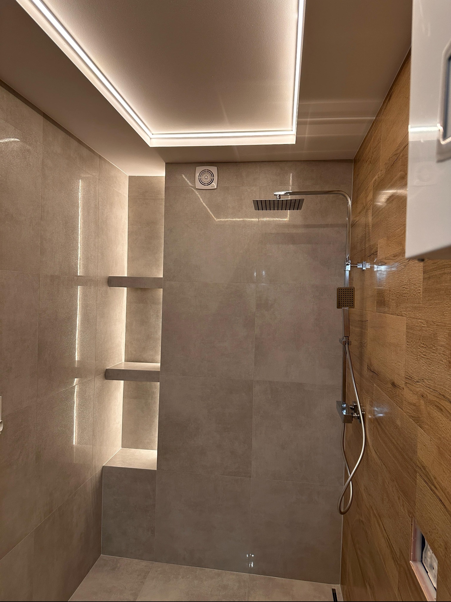 Nowoczesna łazienka po remoncie: szare płytki, drewnopodobne akcenty, podświetlane półki wnękowe i deszczownica. Minimalistyczny design z dbałością o detale.