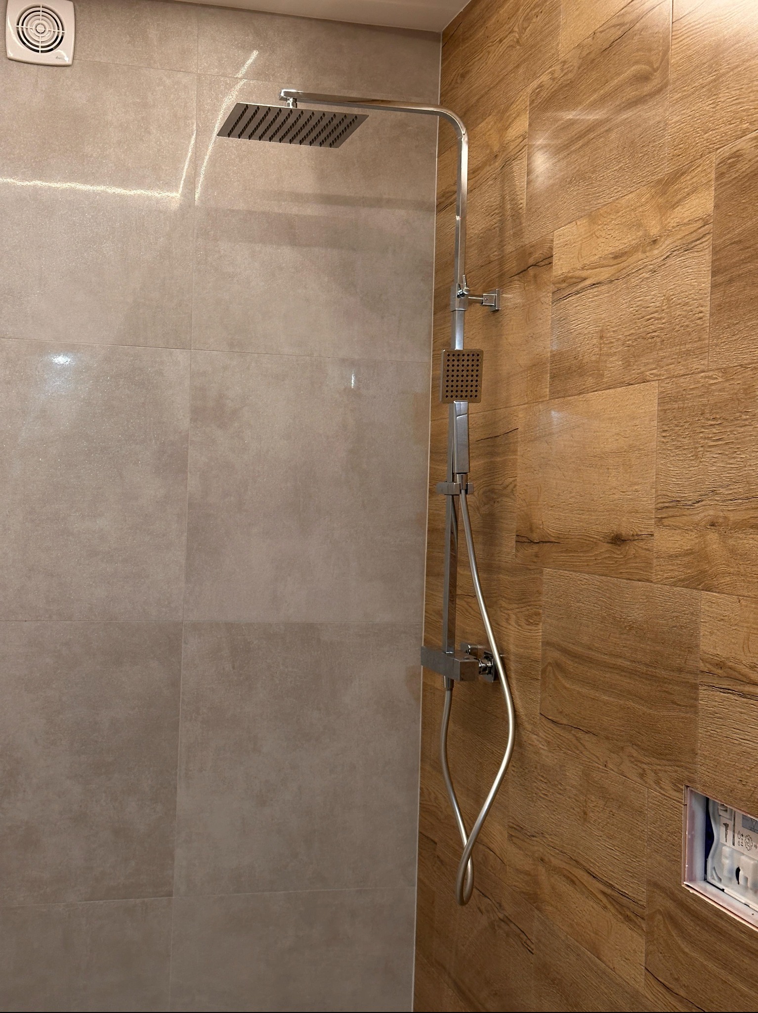 Nowoczesna kabina prysznicowa z deszczownicą i panelami imitującymi drewno, jasne płytki na ścianie po lewej, wentylator w rogu. Minimalistyczny design.