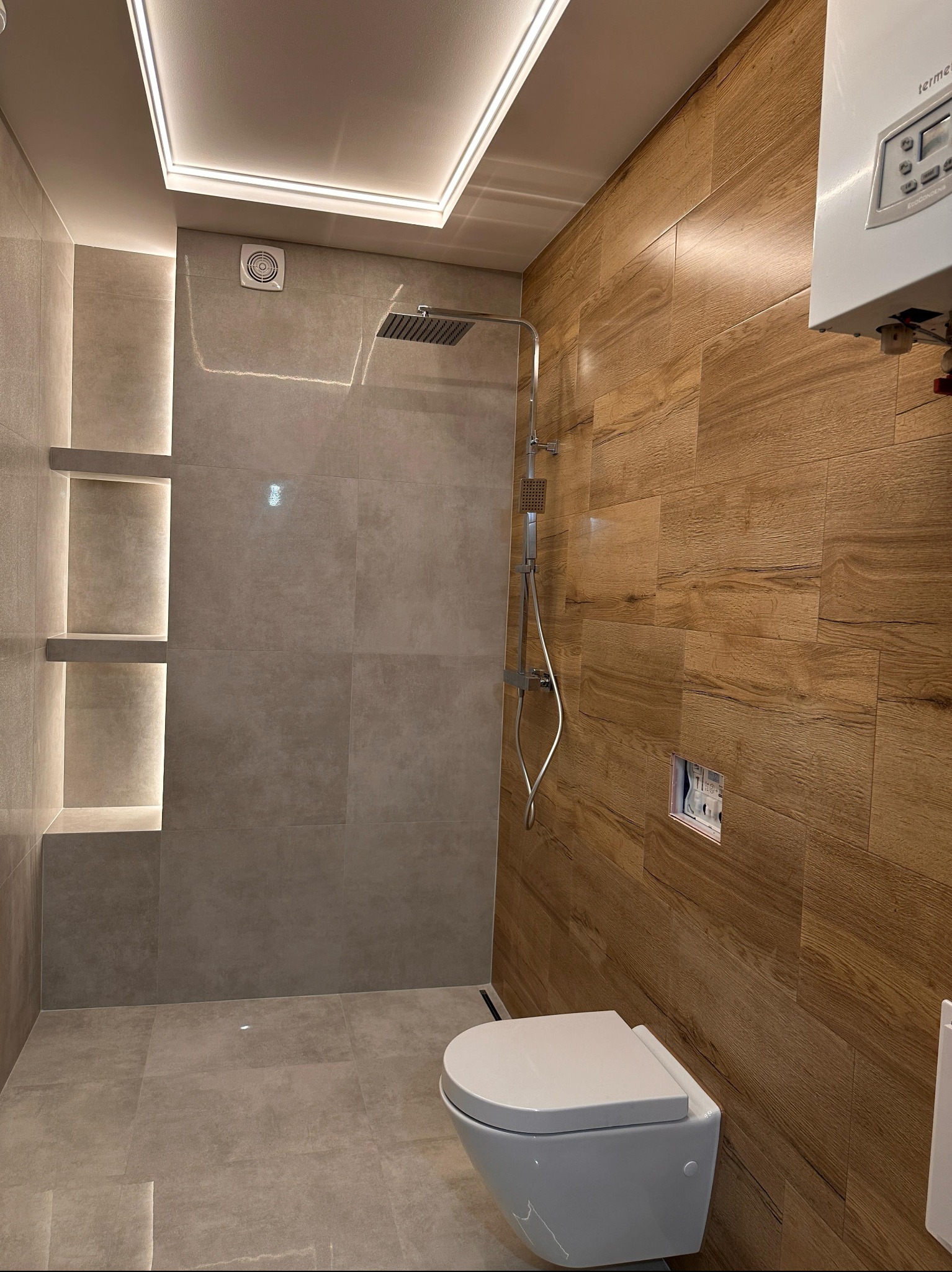 Nowoczesna łazienka z drewnopodobnymi i szarymi płytkami, kabiną prysznicową typu walk-in, podświetlanymi półkami i podwieszaną toaletą. Widoczny bojler.