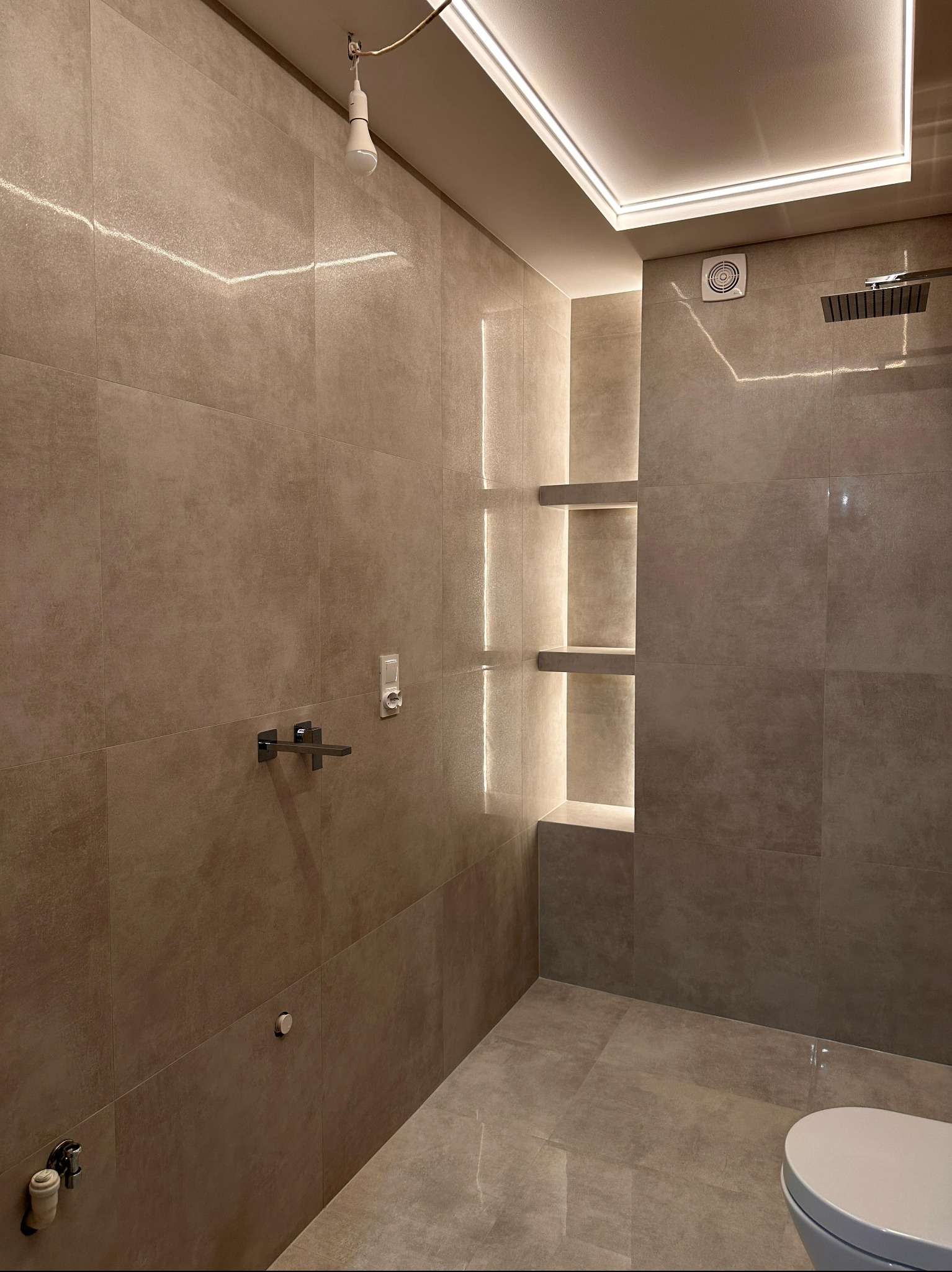 Nowoczesna łazienka w beżowych płytkach z podświetlanymi półkami w ścianie. Widoczna armatura, deszczownica i fragment toalety. Oświetlenie LED na suficie.