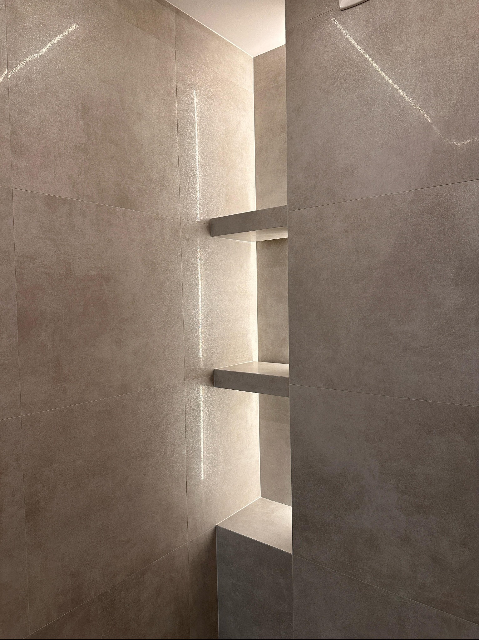 Minimalistyczna łazienka z beżowymi płytkami. Wnęka z półkami podświetlona ledami tworzy nowoczesny akcent. Widoczne fugi i odbicia światła.