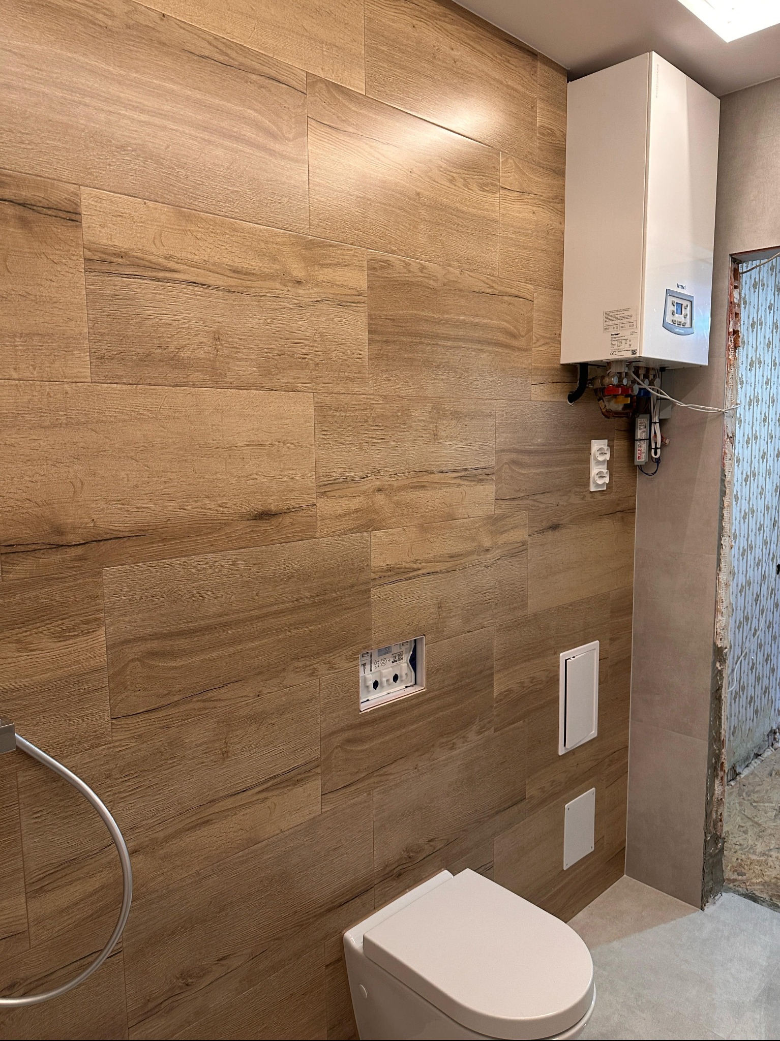 Łazienka w trakcie wykończenia: ściany z płytek imitujących drewno, podwieszana toaleta, bojler na ścianie. Widoczny stan surowy obok.