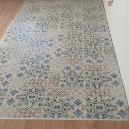 Ułożone płytki podłogowe z wzorem patchworkowym w odcieniach niebieskiego i brązu, widoczne na styku z drewnianą podłogą i białą ścianą, z żółtym statywem w tle.