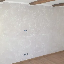 Ściana z dekoracyjną, jasną fakturą przypominającą marmur, z widocznymi drewnianymi belkami stropowymi i otworami po gniazdkach elektrycznych, świeżo położone panele podłogowe.