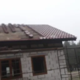 Budowa domu w trakcie układania dachówki ceramicznej; widoczne rusztowania i materiały budowlane.