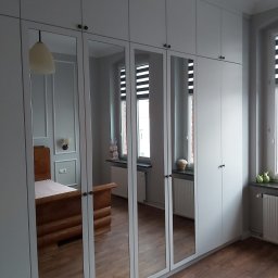 P.P.H.U Awans Marzena Kaczmarek - Biała szafa wnękowa z lustrzanymi drzwiami w sypialni, odbijająca łóżko i okno z żaluzjami.