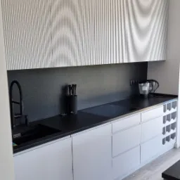 Nowoczesna kuchnia z białymi szafkami z pionowymi żłobieniami, czarnym blatem i czarną armaturą, uzupełniona o drewnopodobną podłogę i półki na wino.