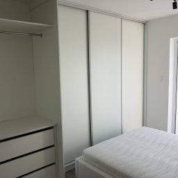 P.P.H.U Awans Marzena Kaczmarek - Nowoczesna szafa wnękowa z przesuwnymi, matowymi drzwiami w minimalistycznej sypialni z białym łóżkiem i oświetleniem szynowym.