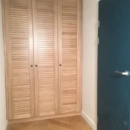 Trzydrzwiowa szafa z jasnego drewna z żaluzjowymi drzwiami, czarnymi gałkami, na tle parkietu ułożonego we wzór jodełki, obok granatowe drzwi z subtelnym wzorem w jodełkę.