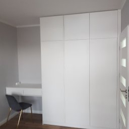 P.P.H.U Awans Marzena Kaczmarek - Biała, minimalistyczna szafa wnękowa z biurkiem w pokoju, krzesło z aksamitnym siedziskiem i złotymi nogami, podłoga z paneli drewnopodobnych, białe drzwi z przeszkleniami.