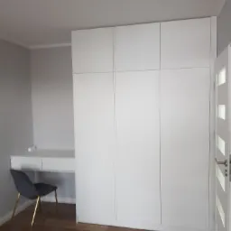 Biała, minimalistyczna szafa wnękowa z biurkiem w pokoju, krzesło z aksamitnym siedziskiem i złotymi nogami, podłoga z paneli drewnopodobnych, białe drzwi z przeszkleniami.