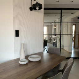 P.P.H.U Awans Marzena Kaczmarek - Jadalnia z drewnianym stołem, szarymi krzesłami i dekoracyjną ścianą z oświetleniem, widok na przeszkloną ścianę działową.