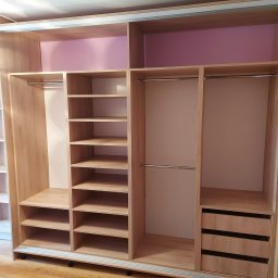 P.P.H.U Awans Marzena Kaczmarek - Wnętrze trzydrzwiowej szafy przesuwnej z jasnego drewna z półkami, drążkami na ubrania i trzema szufladami, widoczne detale wykończenia i mechanizm przesuwny.