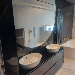 Nowoczesna łazienka z dwoma owalnymi umywalkami nablatowymi na czarnym blacie z marmuru, duże okrągłe lustro z podświetleniem LED, marmurowe ściany w odcieniach czerni i bieli, wanna wolnostojąca...