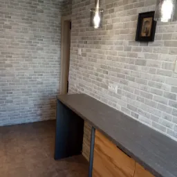 Nowoczesna aranżacja wnętrza z blatem z ciemnego kamienia, drewnianymi szafkami i ścianą z jasnej cegły, oświetlona wiszącymi lampami z przezroczystego szkła.