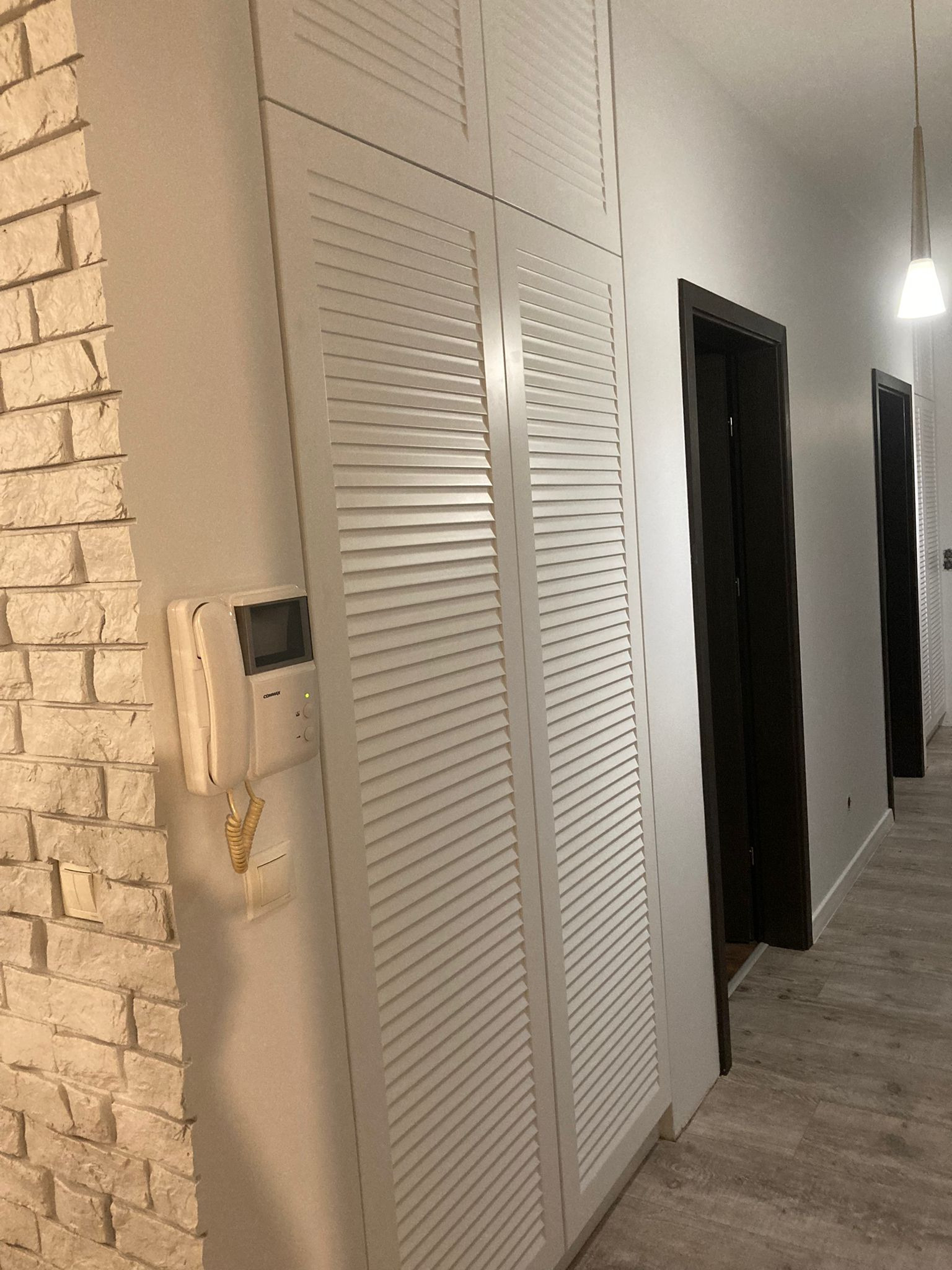 Biała szafa z lamelowymi drzwiami w korytarzu, obok ściana z imitacji cegły i domofon, na podłodze panele imitujące drewno.