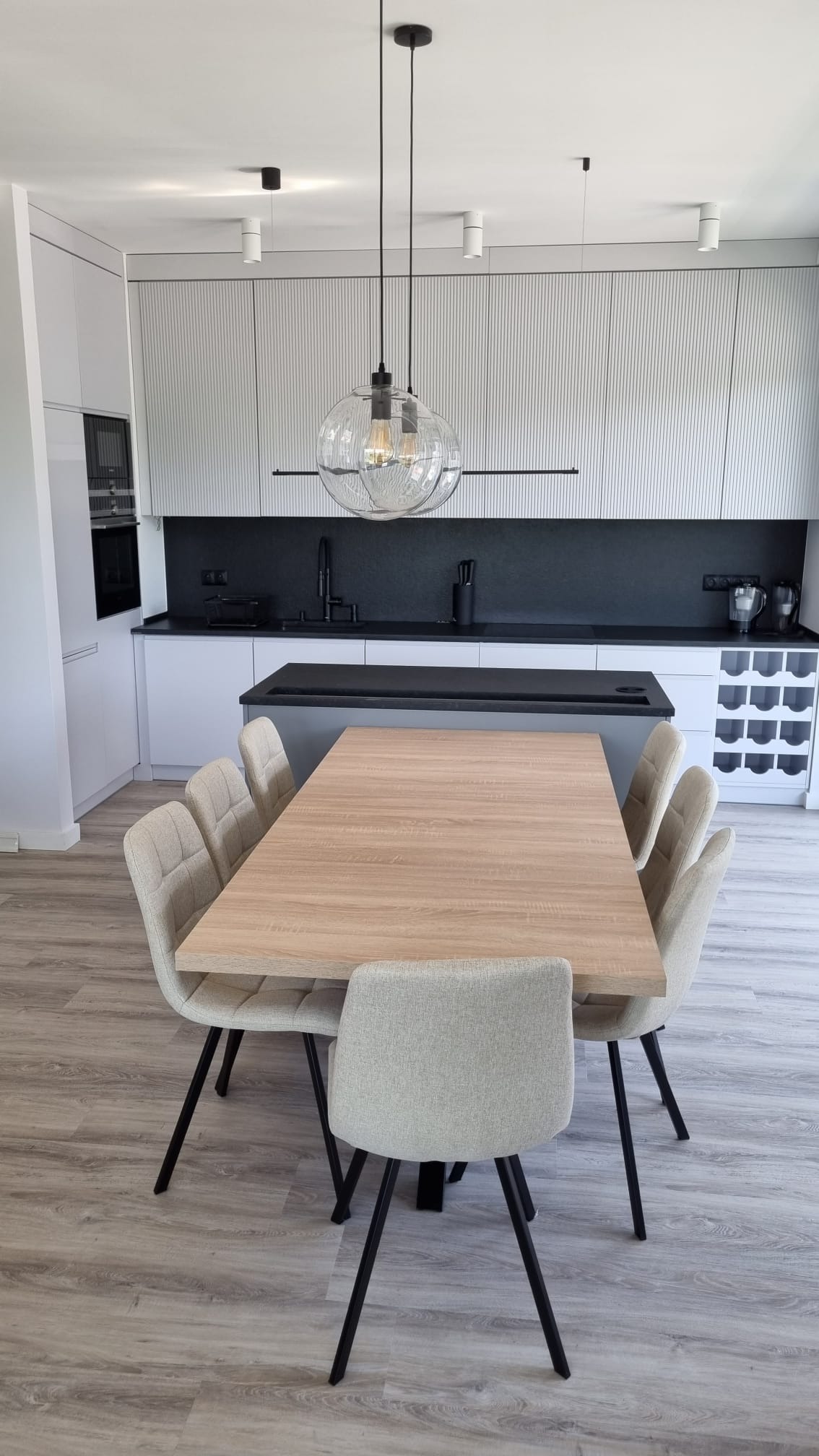 Jasna jadalnia z drewnianym stołem i krzesłami na czarnych nogach, nad stołem szklana lampa wisząca, w tle białe szafki kuchenne z czarnym blatem i wyspą.