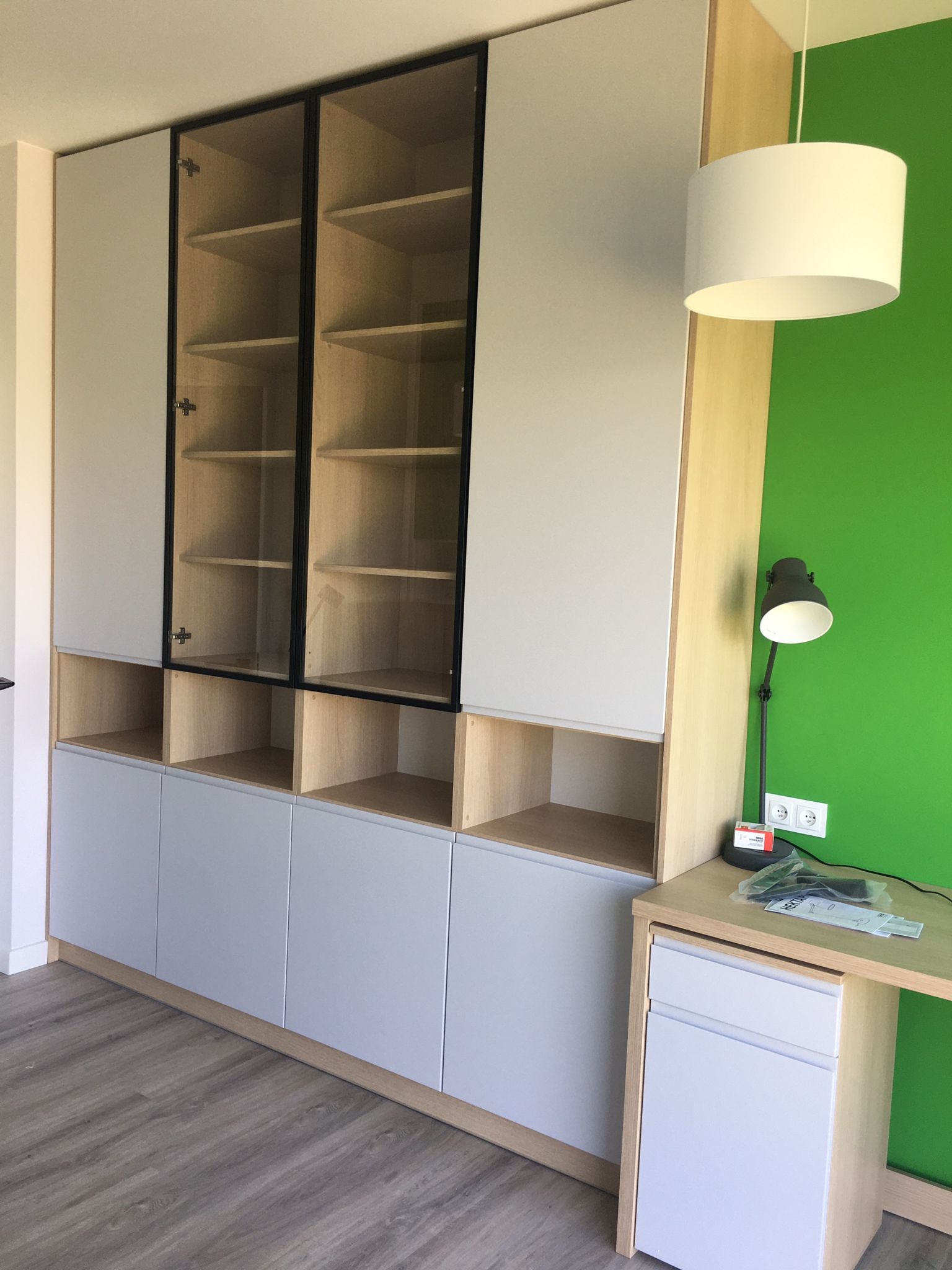 Zabudowa meblowa z szarymi frontami i przeszklonymi witrynami na tle zielonej ściany, uzupełniona biurkiem z szufladami i lampką.