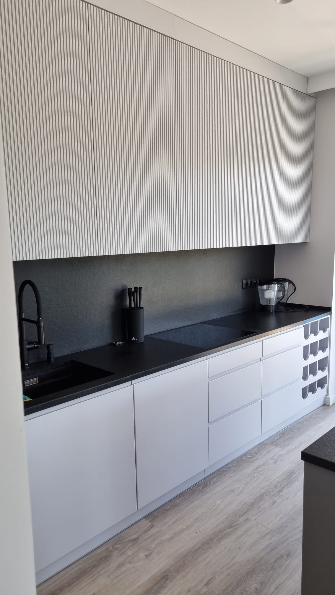 Nowoczesna kuchnia z białymi szafkami z pionowymi żłobieniami, czarnym blatem i czarną armaturą, uzupełniona o drewnopodobną podłogę i półki na wino.