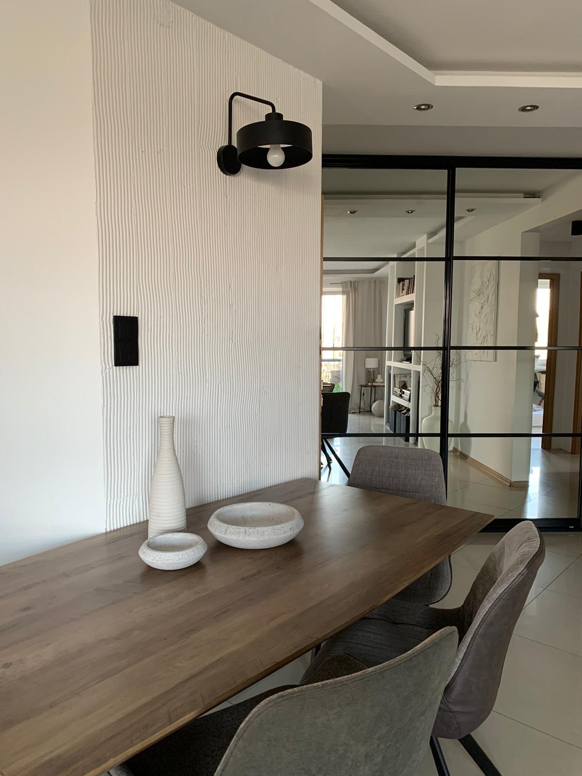 Jadalnia z drewnianym stołem, szarymi krzesłami i dekoracyjną ścianą z oświetleniem, widok na przeszkloną ścianę działową.