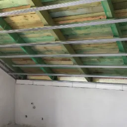 Wnętrze poddasza z widoczną konstrukcją drewnianą impregnowaną na zielono, metalowymi profilami sufitowymi i częściowo ocieploną ścianą szczytową, przygotowane do montażu płyt gipsowo-kartonowych.