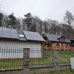 Nowoczesny dom z panelami słonecznymi na dachu, widoczny zza ozdobnego ogrodzenia z kamiennymi słupkami.