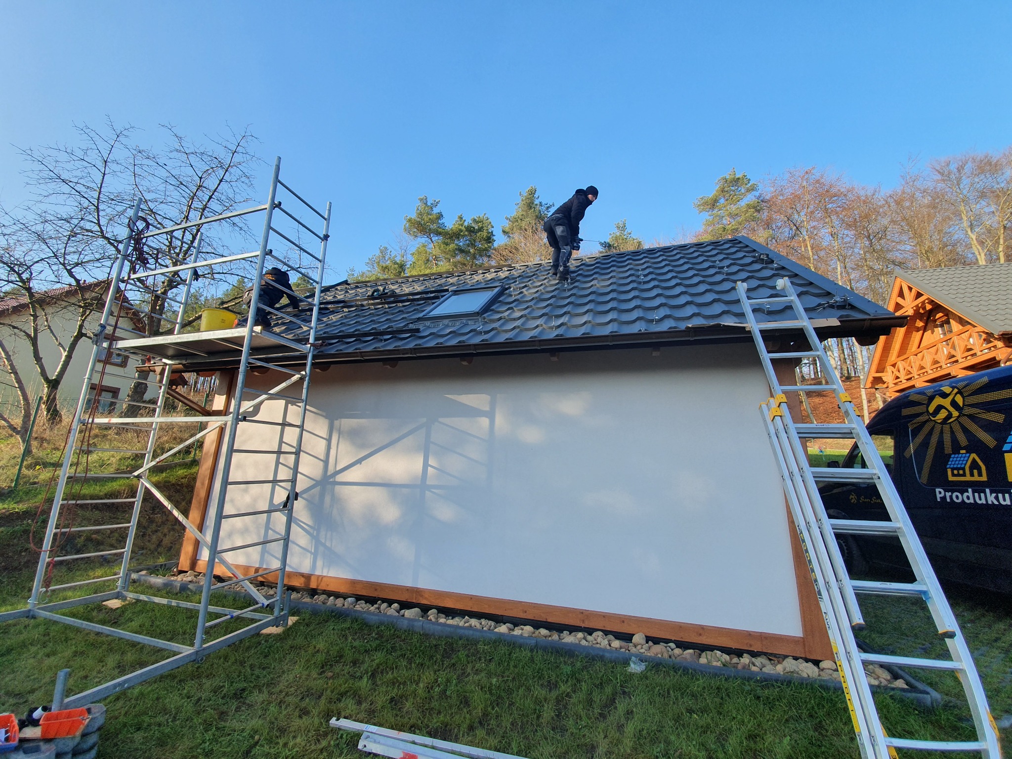 Montaż paneli na dachu budynku z wykorzystaniem rusztowania i drabiny, widoczny samochód firmy z logo.