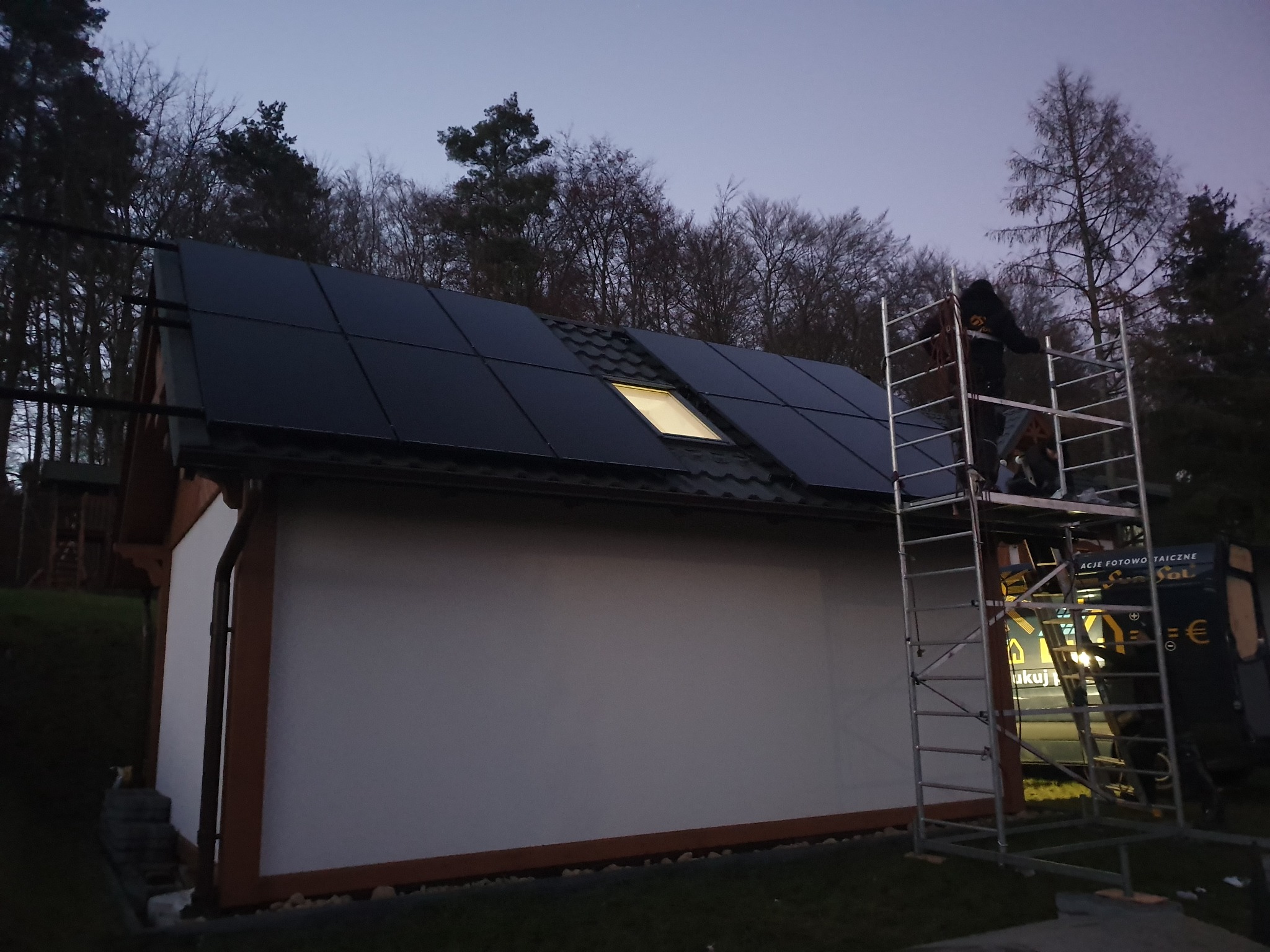 Montaż paneli słonecznych na dachu budynku z oknem dachowym, widoczne rusztowanie i sylwetka osoby na nim, otoczenie leśne w zmierzchu.