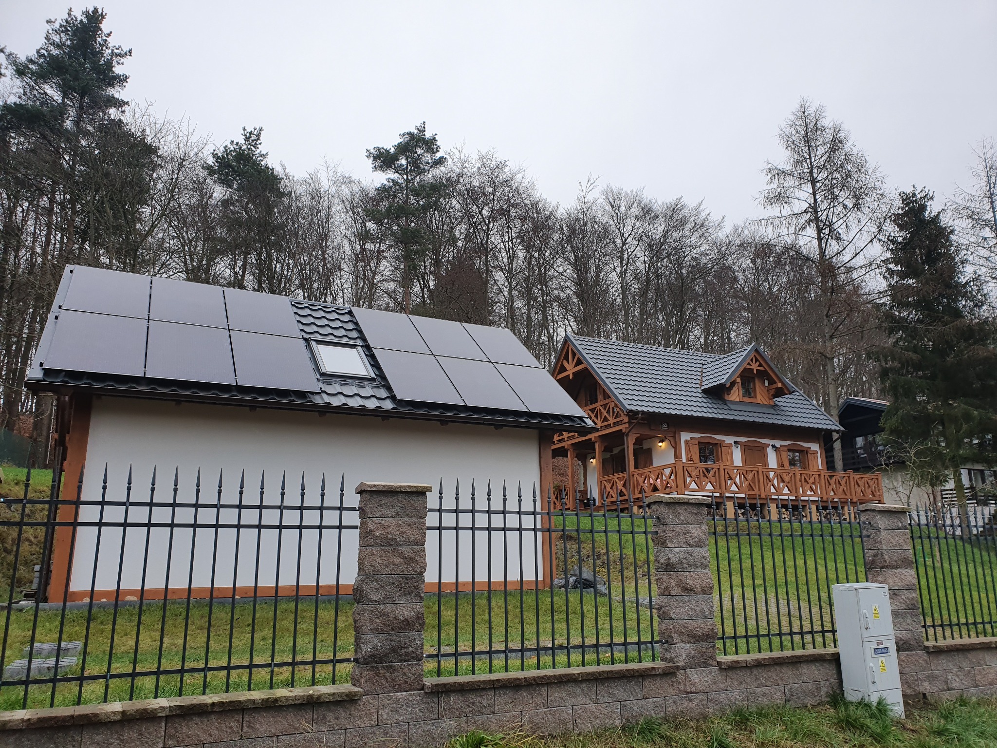 Nowoczesny dom z panelami słonecznymi na dachu, widoczny zza ozdobnego ogrodzenia z kamiennymi słupkami.