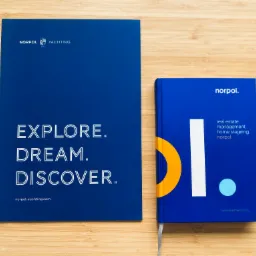 Dwa niebieskie projekty graficzne firmy Norpol na drewnianym tle: broszura z hasłem 'EXPLORE. DREAM. DISCOVER.' i notes z abstrakcyjnym logo.