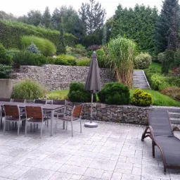 Rozległy taras z kamienną posadzką, otoczony bujną zielenią, w tle skarpa umocniona gabionami, schody prowadzące do ogrodu, zestaw mebli ogrodowych i leżak.