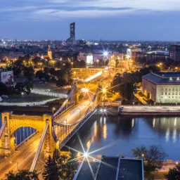 Nocny widok na Wrocław z mostem Grunwaldzkim, Odrą i oświetlonym budynkiem uniwersytetu, z widocznymi smugami świateł samochodów i wieżowcem w tle.