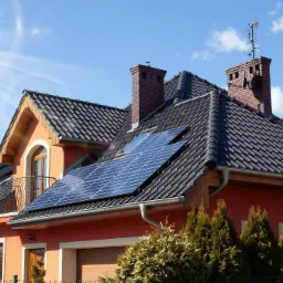 Pomarańczowy dom z ciemną dachówką, wyposażony w panele słoneczne na dachu, z widocznym kominem i anteną satelitarną. Na pierwszym planie iglaki.