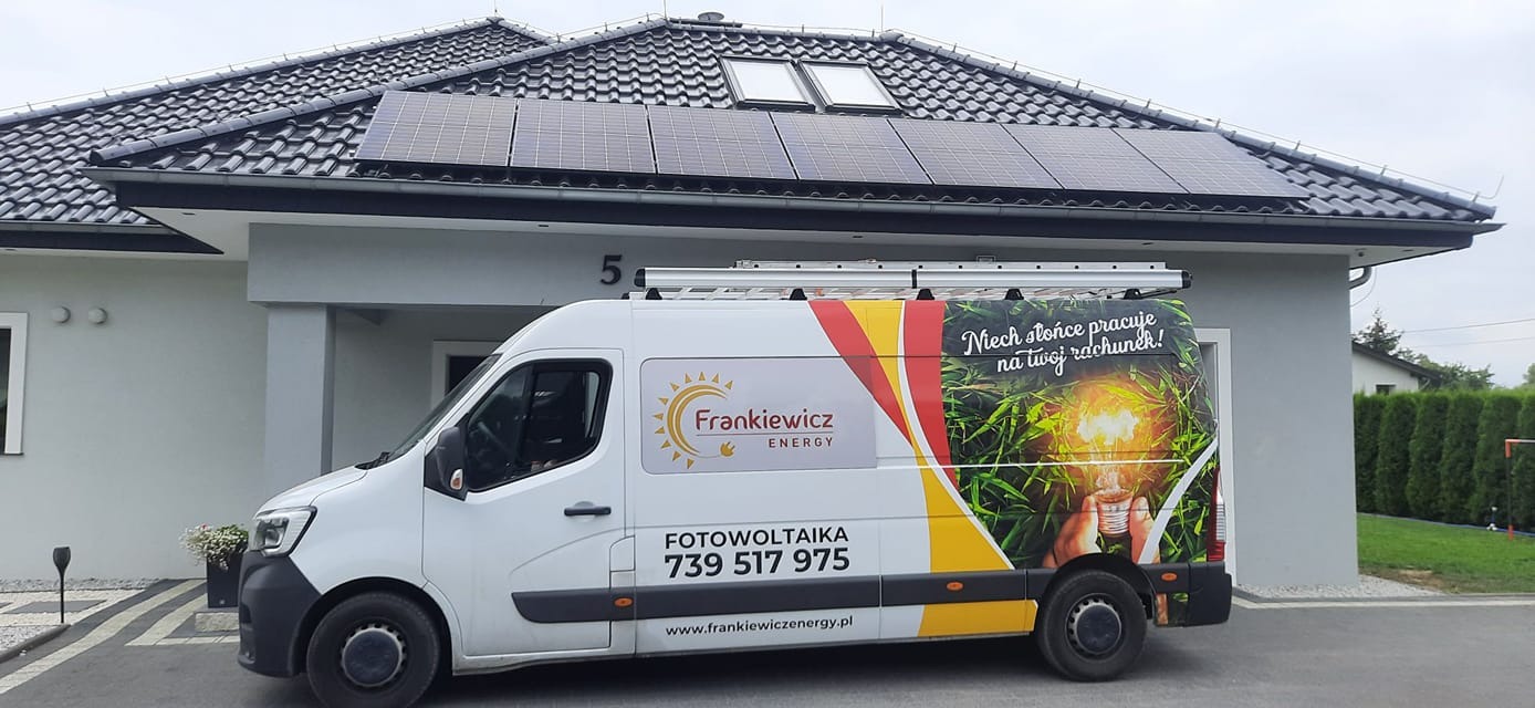 Panele fotowoltaiczne na dachu domu jednorodzinnego z szarą elewacją, obok zaparkowany biały samochód dostawczy z reklamą firmy fotowoltaicznej, widoczne okna dachowe i drabina na dachu samochodu.