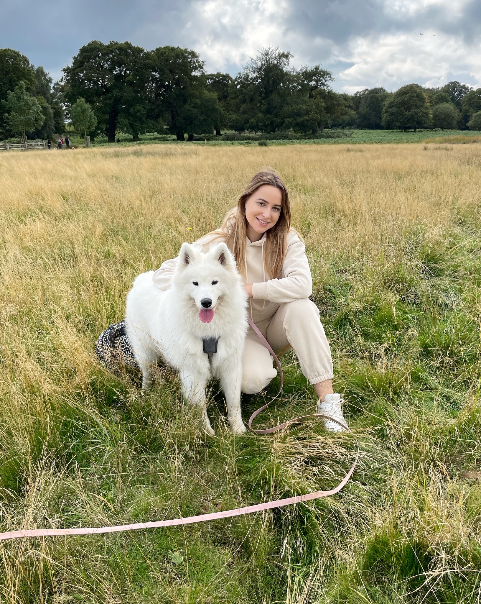 Uśmiechnięta kobieta w beżowym dresie kuca obok białego psa rasy Samoyed w szelkach na łące z wysoką trawą, różowa smycz leży na ziemi.