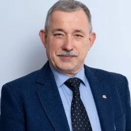 Portret uśmiechniętego mężczyzny w niebieskiej marynarce i krawacie w kropki, z przypinką w klapie.