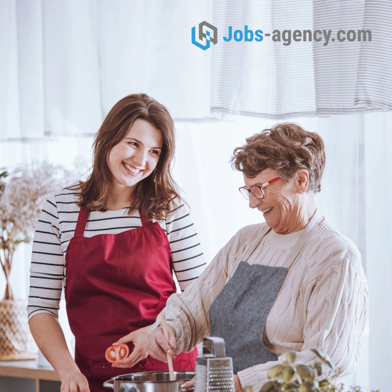 Uśmiechnięta młoda kobieta w czerwonym fartuchu trzyma połówkę pomidora, podczas gdy starsza pani w szarym fartuchu miesza w garnku, obydwie przygotowują posiłek w jasnej kuchni