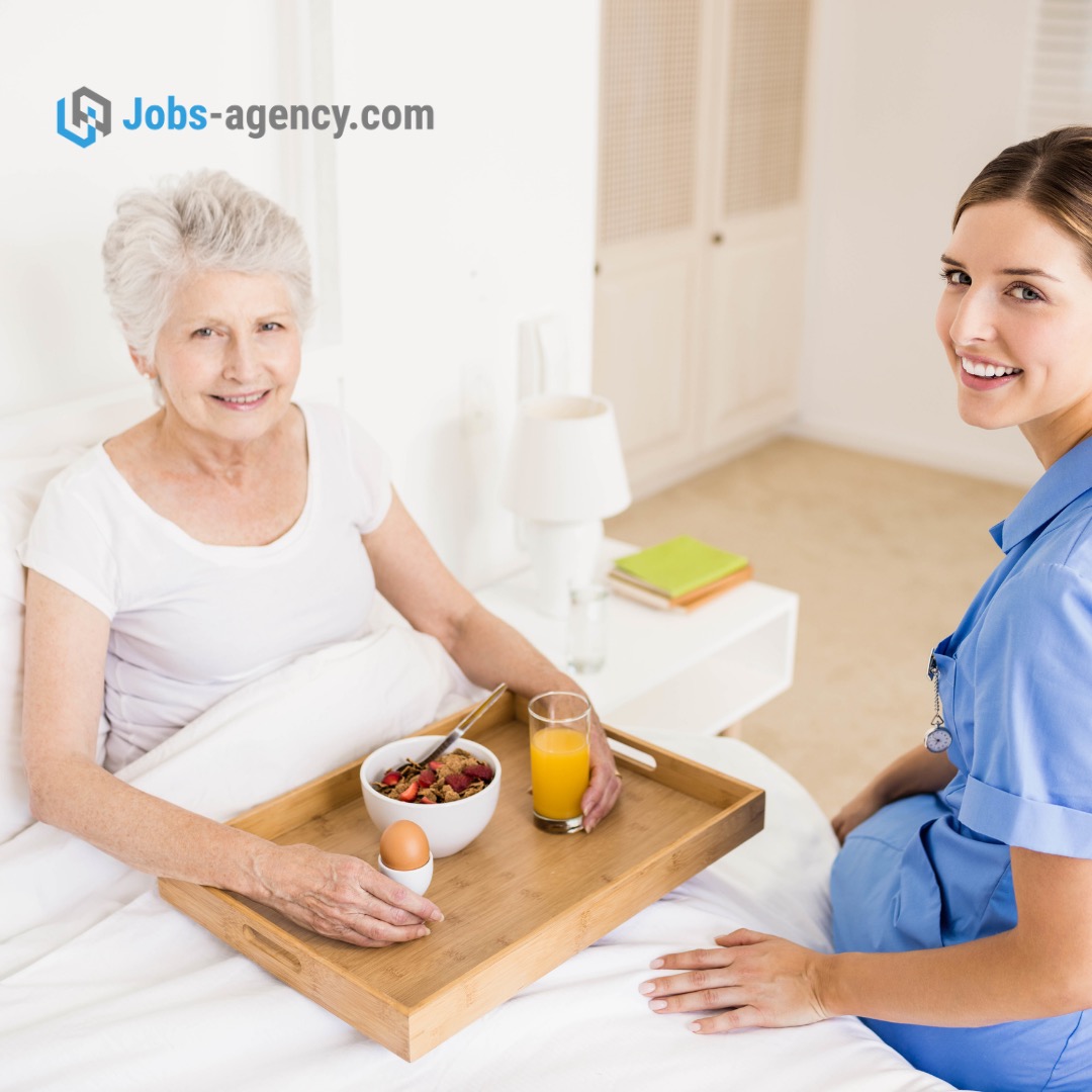 Uśmiechnięta starsza kobieta w łóżku, trzymająca na drewnianej tacy śniadanie: miska z płatkami i owocami, jajko na miękko, szklanka soku pomarańczowego; obok uśmiechnięta opiekunka w niebieskim...