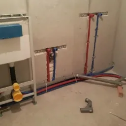 Stelaż podtynkowy WC z widocznymi przyłączami wodnymi i kanalizacyjnymi w surowym stanie, czerwone i niebieskie rury prowadzone w ścianie, szare rury kanalizacyjne na podłodze, cegła podpierająca...