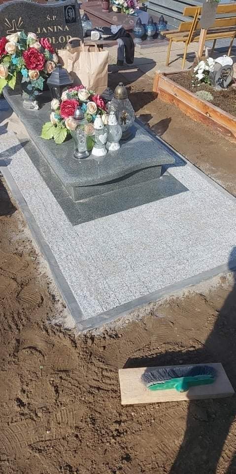 Granitowy nagrobek z kwiatami i zniczami, otoczony świeżo ułożoną kostką granitową, widoczna szczotka do czyszczenia na ziemi.