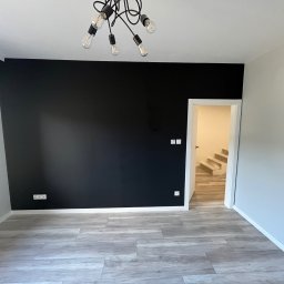 Piotr Piórkowski - Pomalowane na czarno ściana w nowym mieszkaniu, jasnoszare panele podłogowe imitujące drewno, nowoczesna lampa z żarówkami Edisona, widok na schody w głębi.