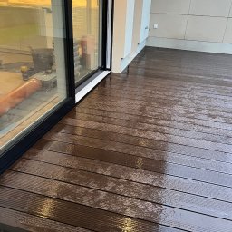 Piotr Piórkowski - Nowoczesny taras z desek kompozytowych w odcieniach brązu, częściowo mokry, przylegający do budynku z dużym oknem. Widoczne narzędzia i materiały wewnątrz.