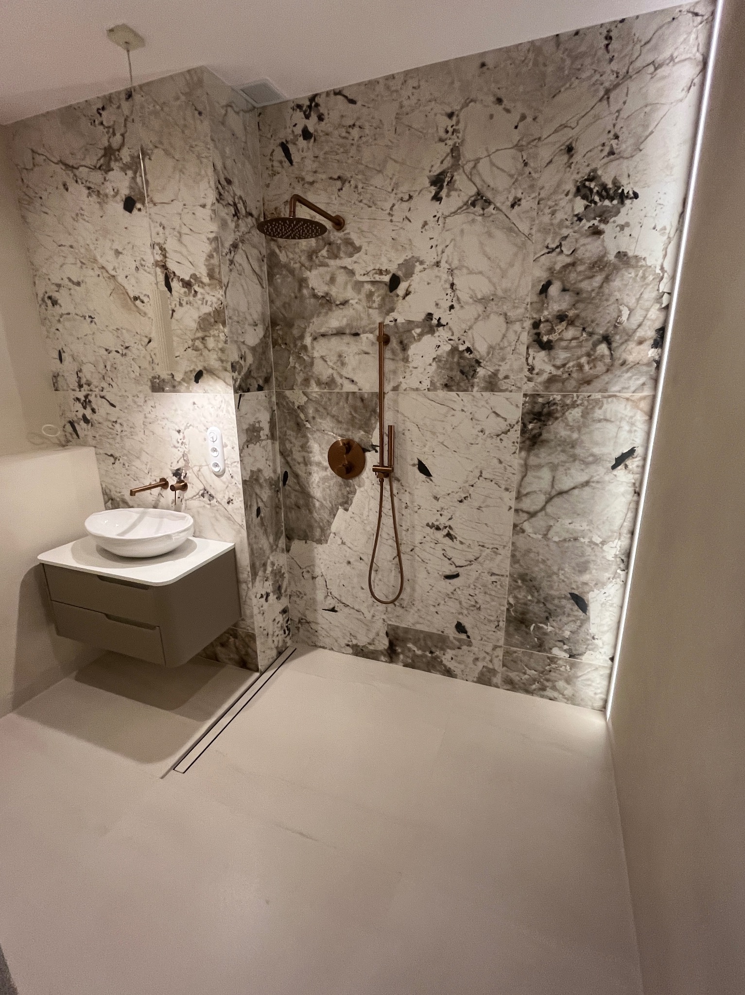 Nowoczesna łazienka z marmurowymi płytkami na ścianach, deszczownicą w kolorze miedzi, umywalką nablatową na szafce z szufladami i odpływem liniowym w podłodze.