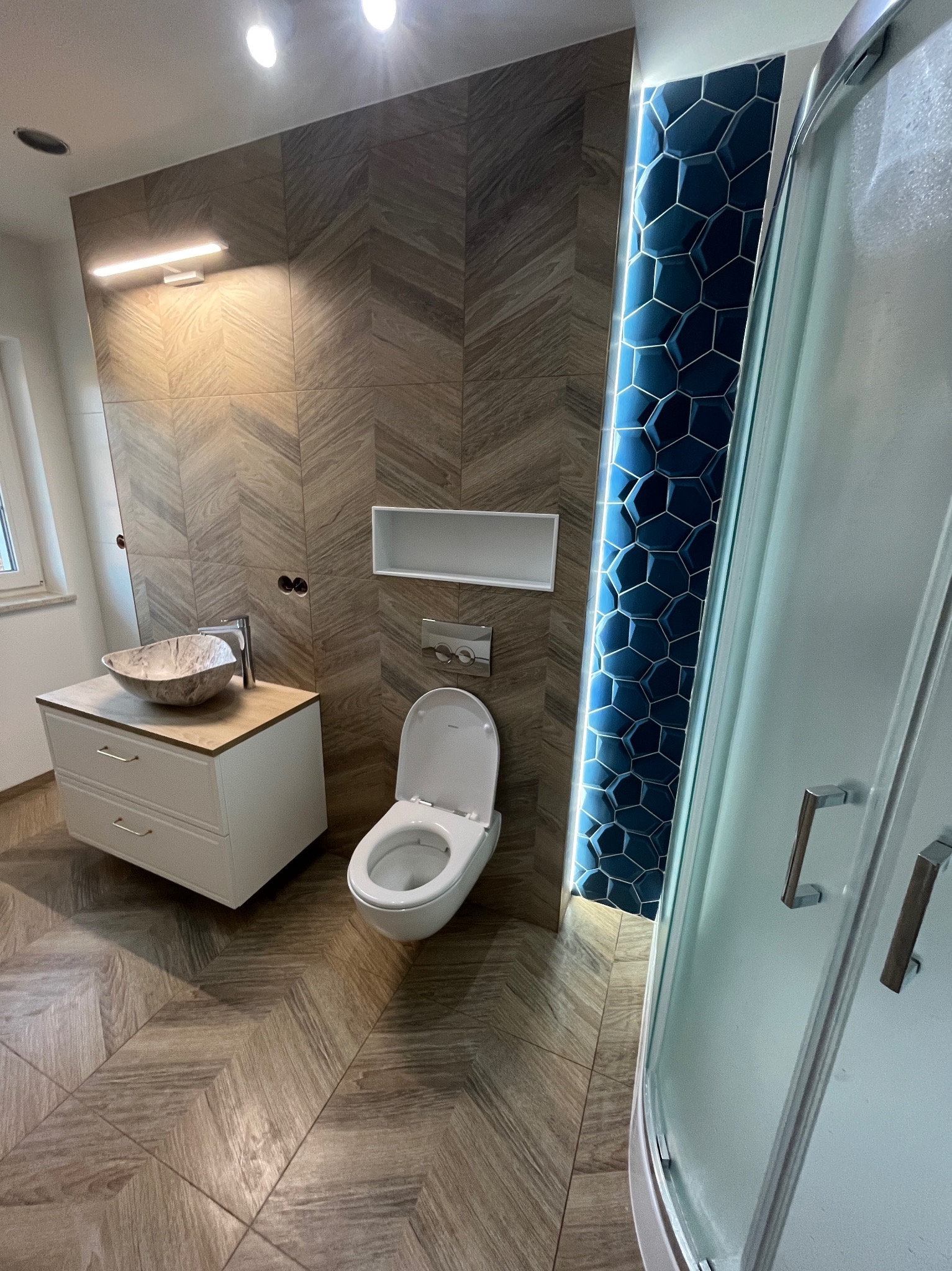 Nowoczesna łazienka z drewnopodobnymi płytkami ułożonymi w jodełkę na podłodze i ścianach, marmurowa umywalka nablatowa, toaleta oraz kabina prysznicowa z niebieskimi, heksagonalnymi płytkami...