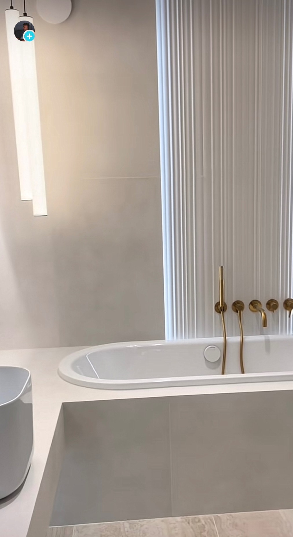 Minimalistyczna łazienka w odcieniach bieli i szarości z owalną wanną, złotą armaturą i dekoracyjną ścianą z pionowymi żłobieniami.