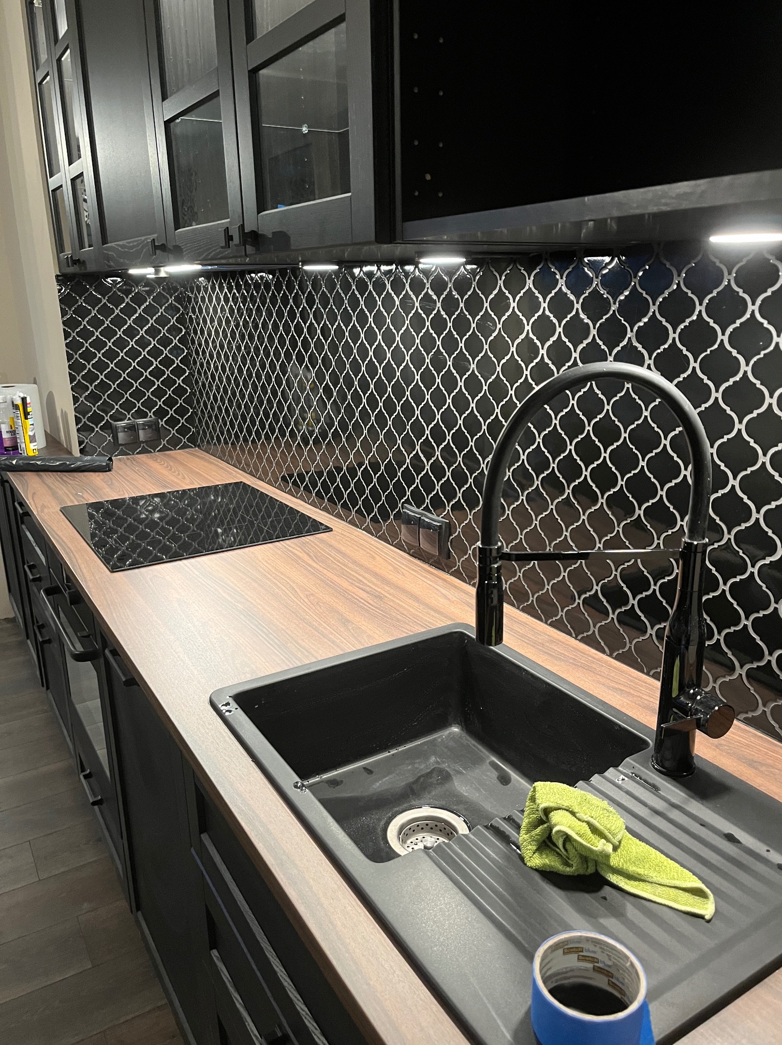 Czarny zlew z ociekaczem, bateria w kolorze czarnym, zielona ściereczka i niebieska taśma malarska na blacie z drewnopodobnym wykończeniem, nad nim czarna płyta indukcyjna i mozaika w czarno-białą...