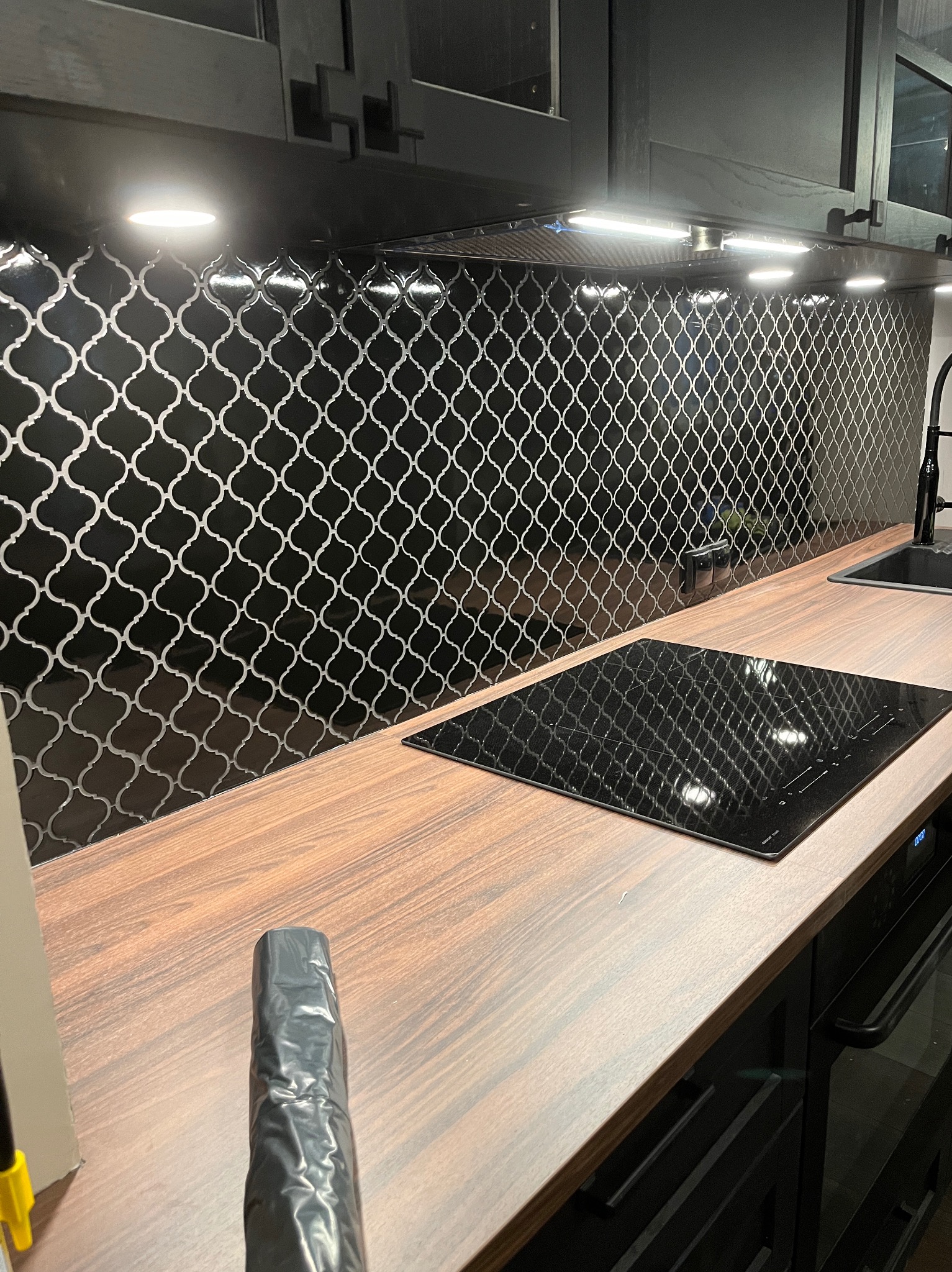 Czarna mozaika z białym wzorem marokańskim nad drewnianym blatem kuchennym z płytą indukcyjną i czarnymi szafkami.