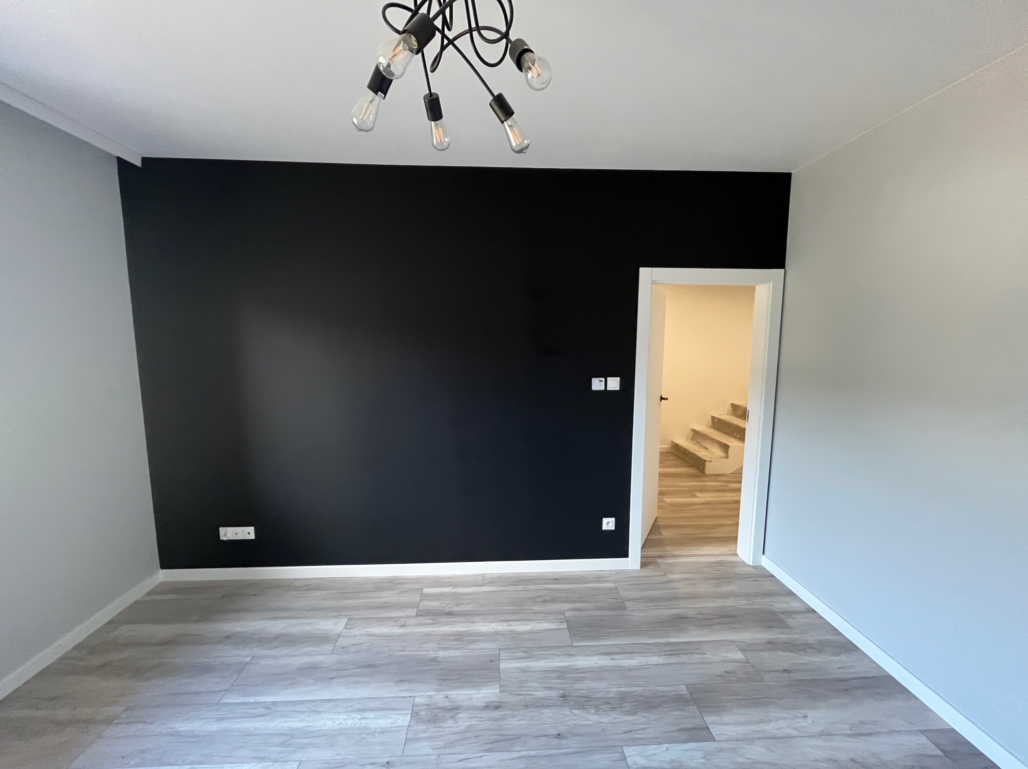 Pomalowane na czarno ściana w nowym mieszkaniu, jasnoszare panele podłogowe imitujące drewno, nowoczesna lampa z żarówkami Edisona, widok na schody w głębi.
